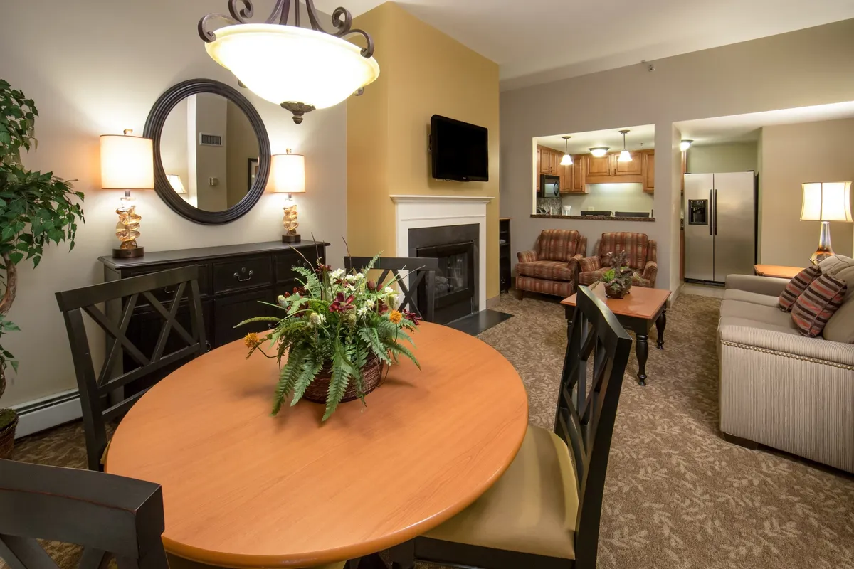 This image features a cozy interior living space with a fireplace as the focal point. The room includes a comfortable seating area, a dining table with a floral centerpiece, and a wall-mounted television—offering a clear sense of the villa’s main living and gathering area. The warm lighting and inviting decor suggest a space perfect for relaxation, family dinners, or socializing by the fire.