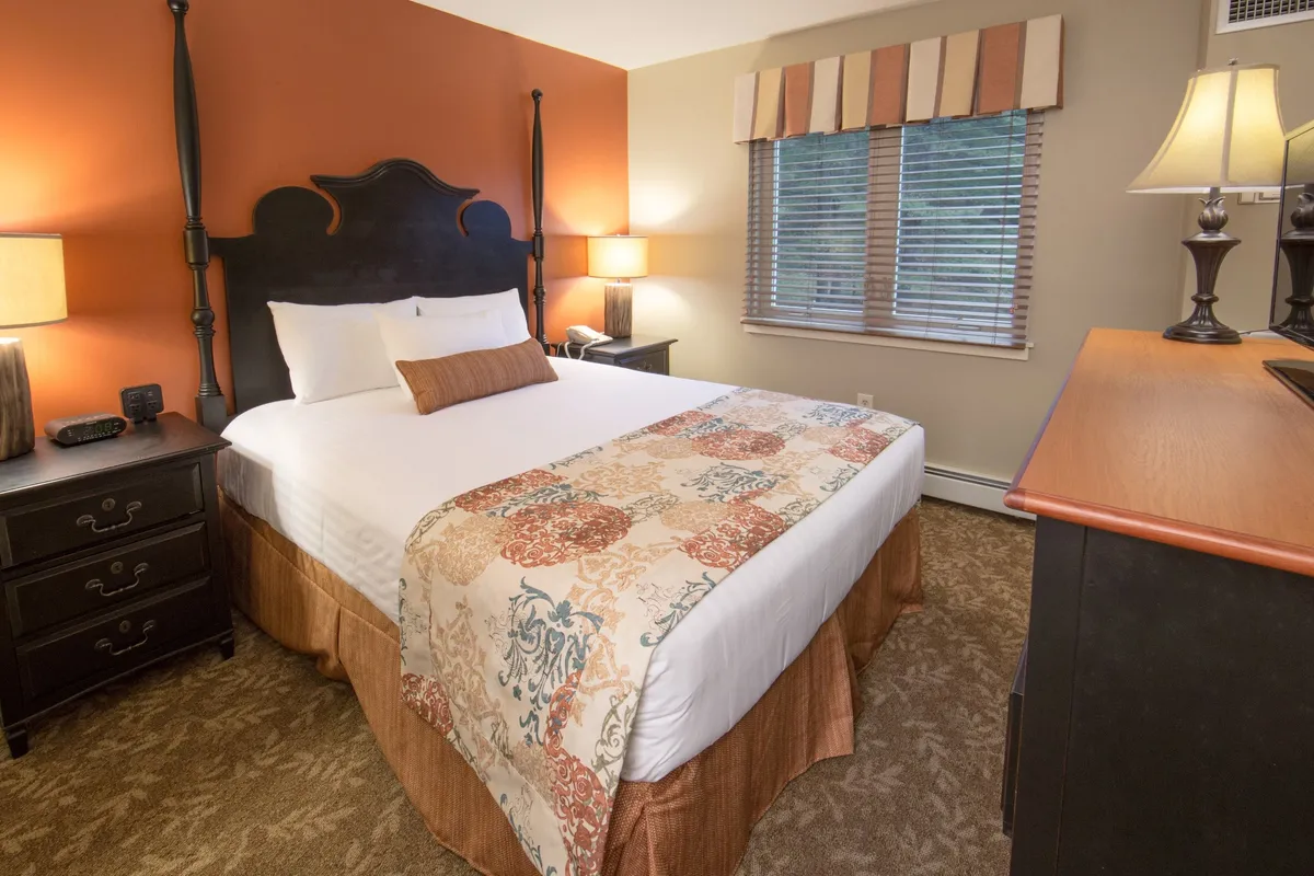 A peaceful bedroom scene featuring a canopy bed adorned with decorative pillows and nightstands with lamps. A window with blinds lets in natural light, creating an atmosphere ideal for relaxing, reading, or watching television.