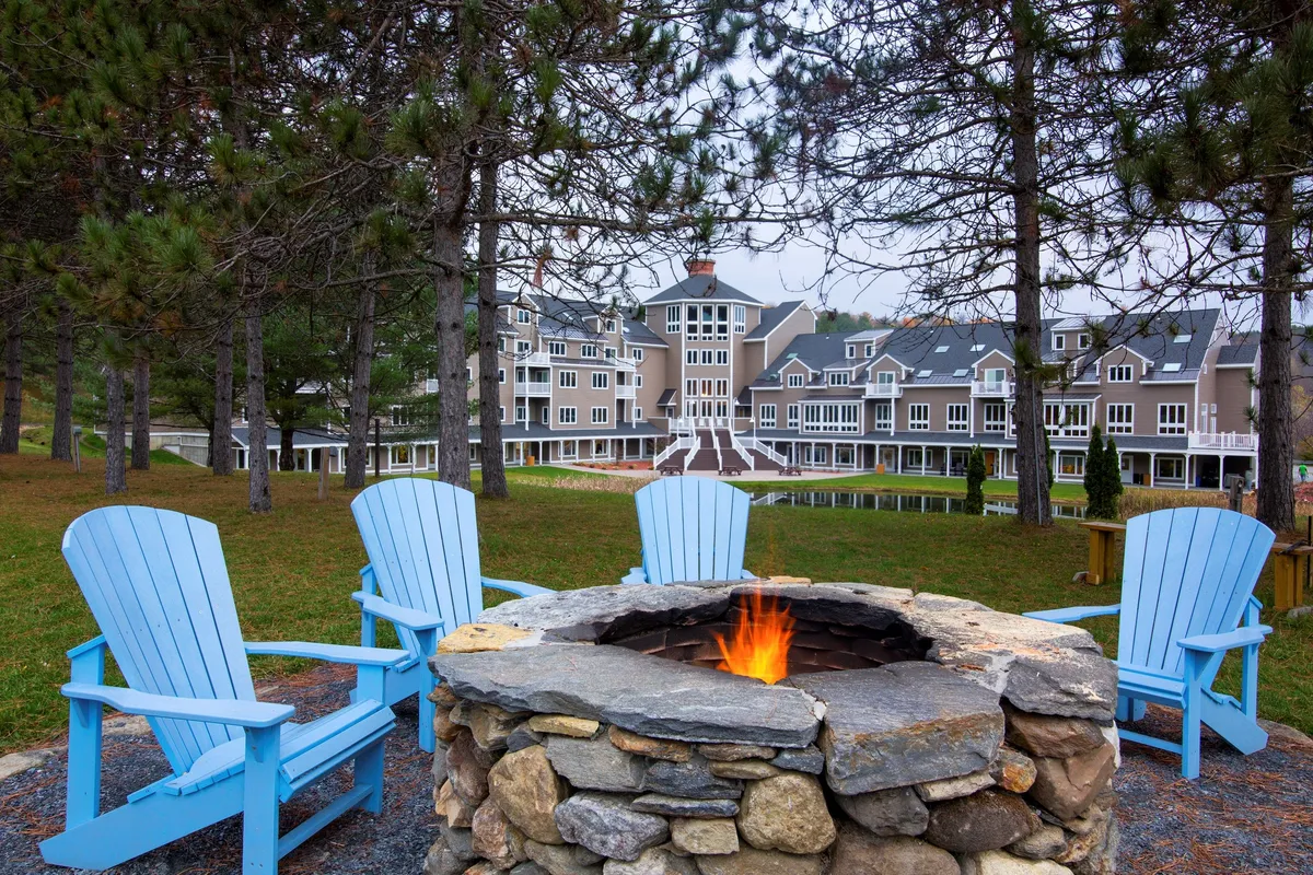 The communal fire pit area is framed by blue Adirondack chairs and the resort building in the background. Guests gather around the fire, enjoying the outdoors in a cozy, social atmosphere—perfect for unwinding after an active day.