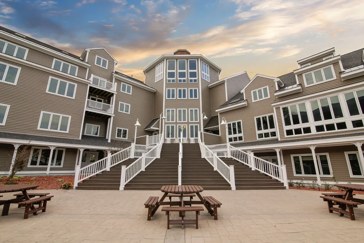 A vibrant exterior shot of the resort's grand entrance, featuring a wide staircase, large windows, and comfortable outdoor seating. The setting is lively with guests arriving and mingling, illuminated by the warm glow of sunset. This image conveys a sense of welcome and excitement, hinting at the communal and family-centered atmosphere within the resort.