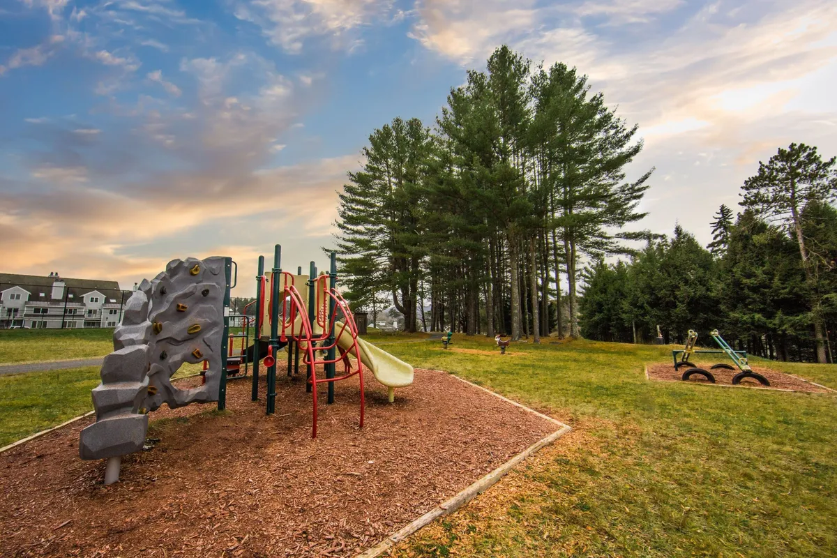 A cheerful scene of a well-maintained playground surrounded by trees and open grassy areas. Children are seen playing on the equipment while families gather nearby, some picnicking or relaxing. The image is full of life, showcasing the outdoor recreation opportunities and family-friendly amenities the resort offers.