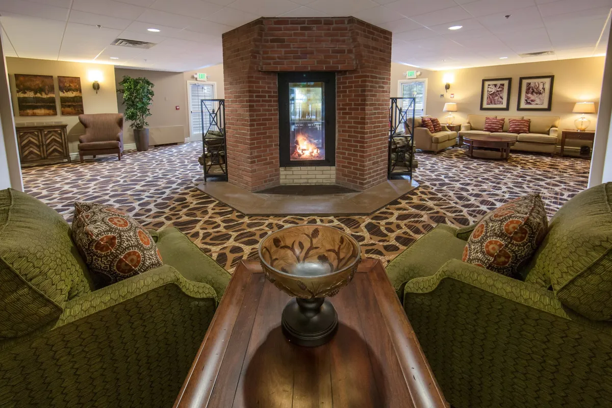 A stylish living room centered around a crackling fireplace, surrounded by green sofas and a decorative bowl on the coffee table. The inviting ambiance is enhanced by warm lighting, making it an ideal spot for socializing, reading, or simply relaxing by the fire.